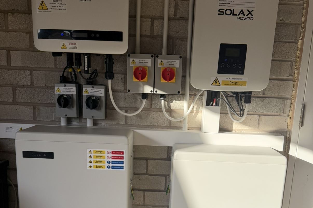 An existing solar inverter with a separate battery inverter and batteries fitted alongside, on a garage wall in St Helens